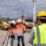 Two construction engineers in hard hats and high-visibility jackets review a set of blueprints next to a surveying transit level at an active outdoor construction site.