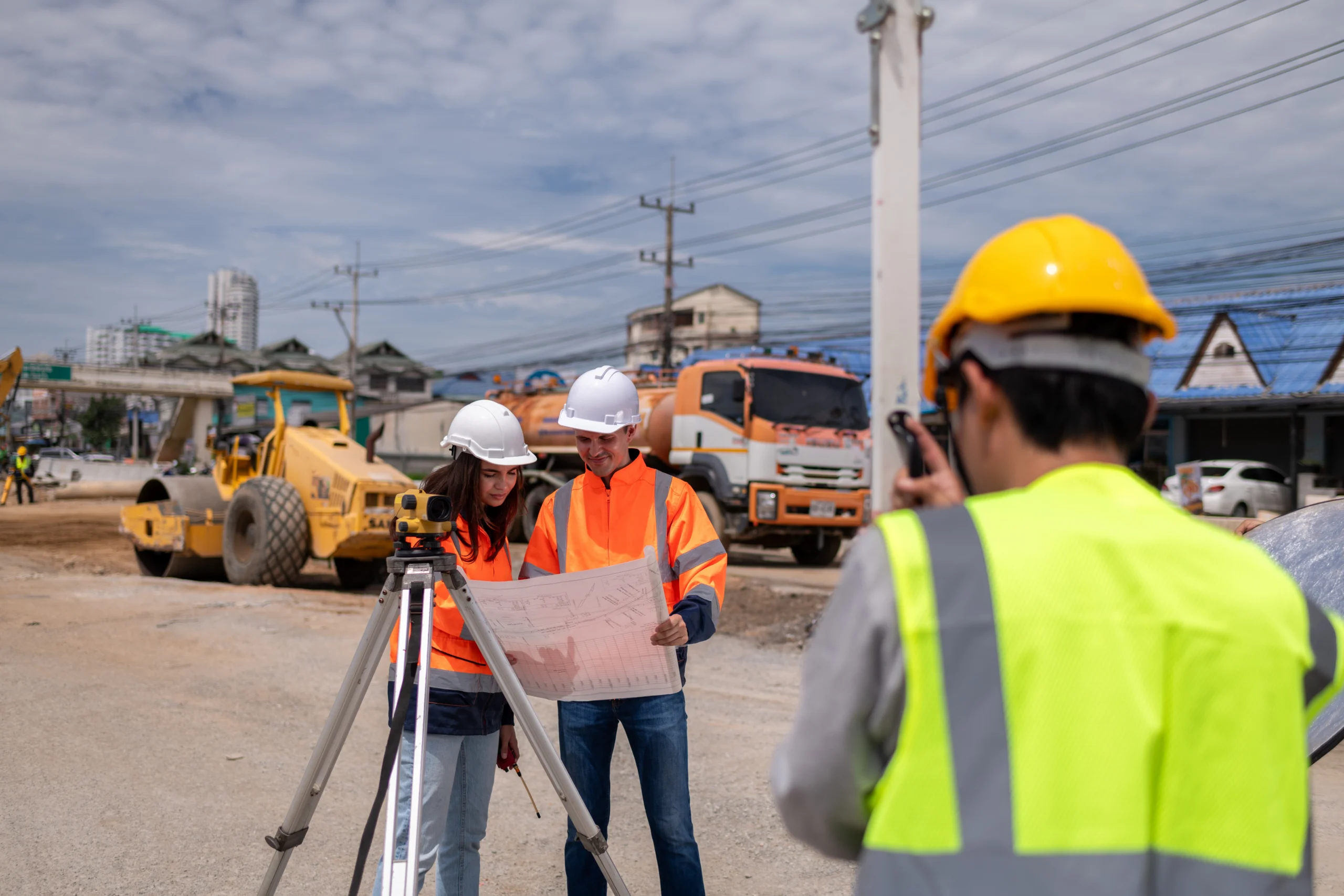 Two construction engineers in hard hats and high-visibility jackets review a set of blueprints next to a surveying transit level at an active outdoor construction site.