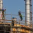 Three construction workers wearing safety harnesses and blue hard hats assemble metal scaffolding outside an industrial processing facility.