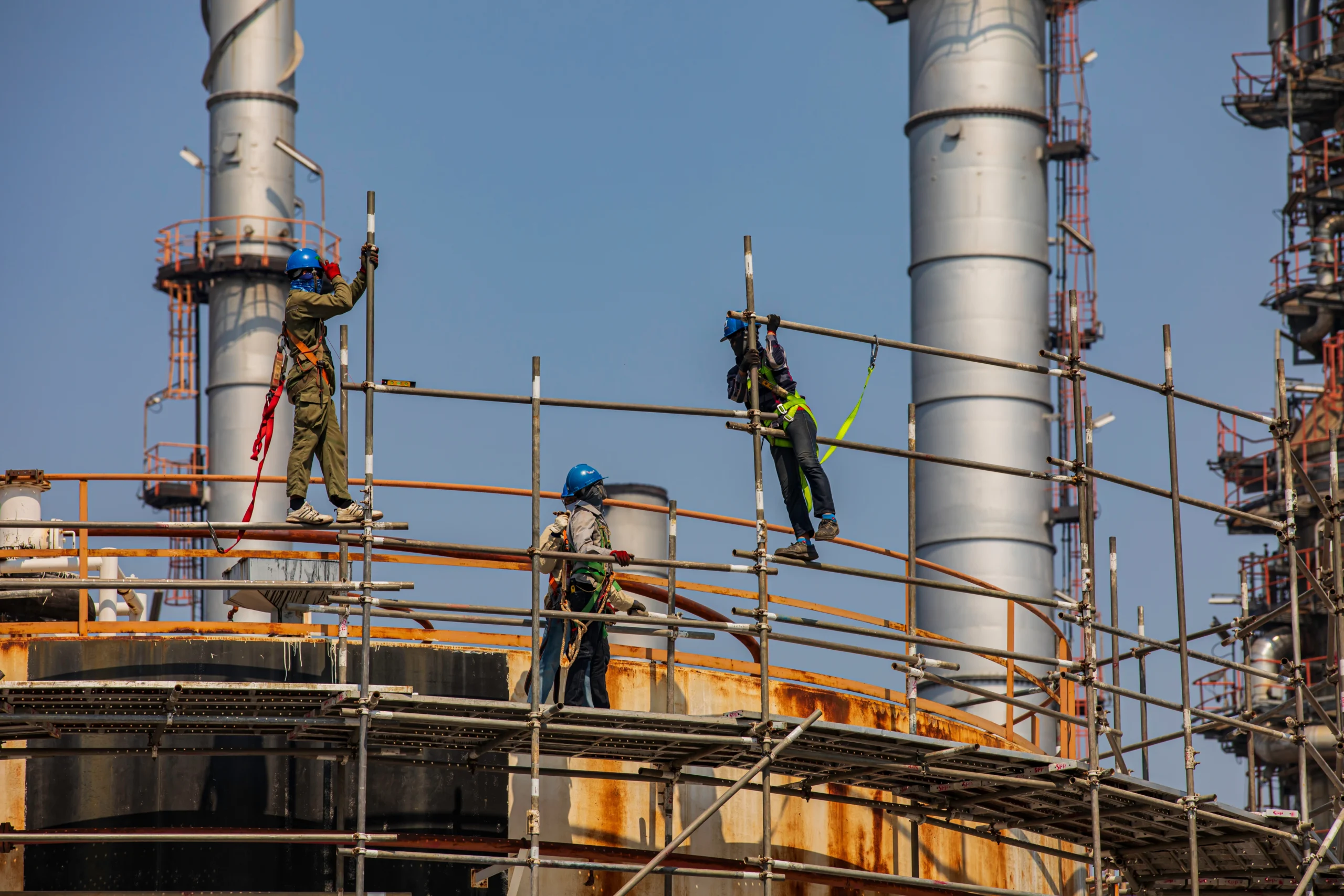 Three construction workers wearing safety harnesses and blue hard hats assemble metal scaffolding outside an industrial processing facility.