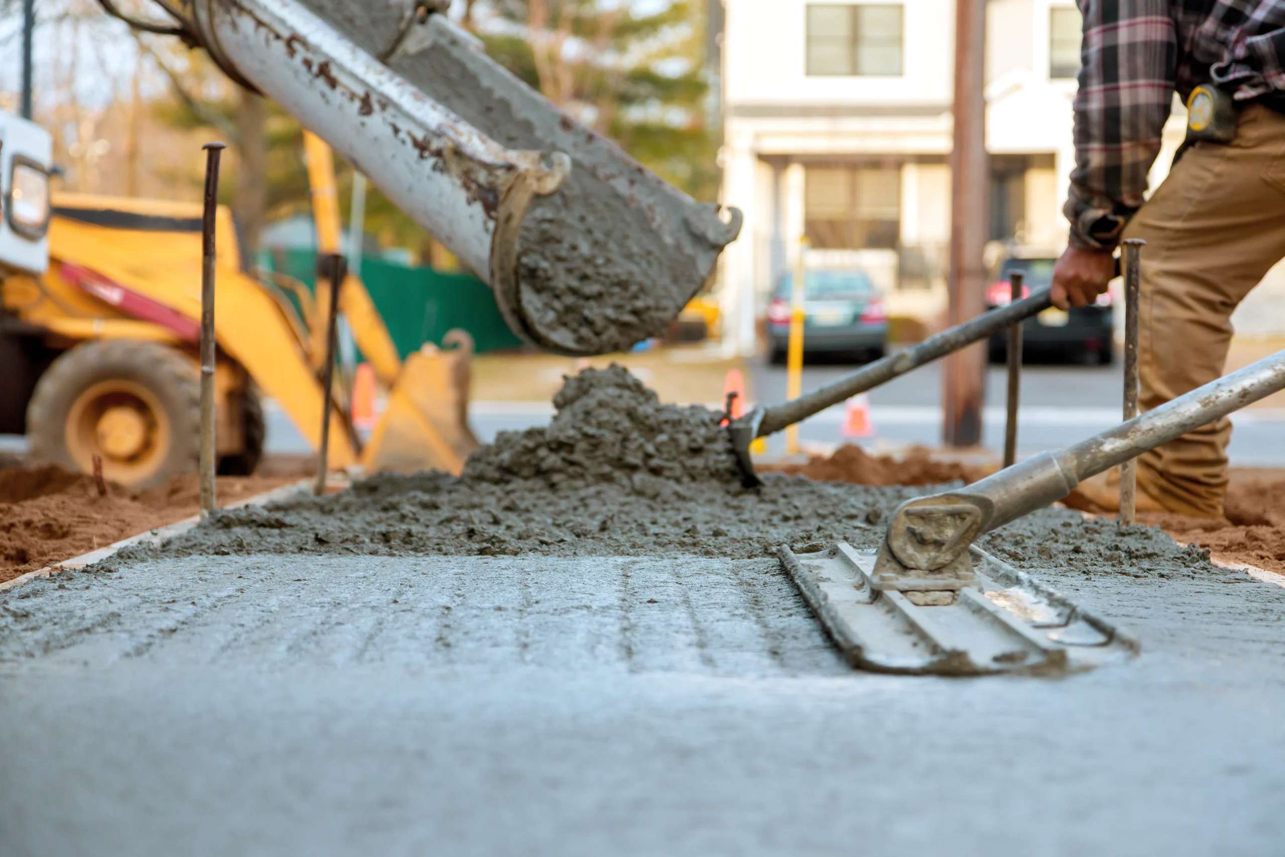 Construction worker pouring and leveling concrete at a worksite, with a concrete mixer and construction equipment in the background.